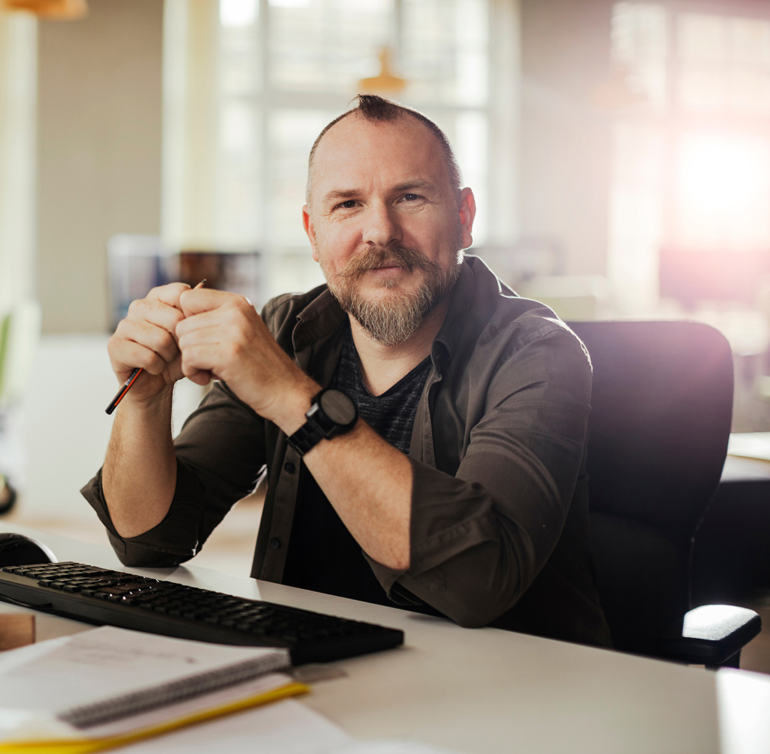 Homem com barba sentado em uma mesa de escritório, sorrindo para a câmera, com as mãos cruzadas sobre um lápis e um relógio no pulso, em um ambiente de escritório com iluminação natural ao fundo.