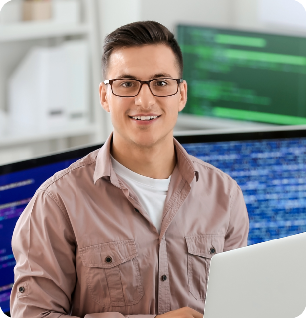Homem jovem com óculos, sorrindo e olhando para a câmera, vestindo uma camisa casual bege, sentado em frente a um laptop com telas de código ao fundo em um ambiente de escritório.
